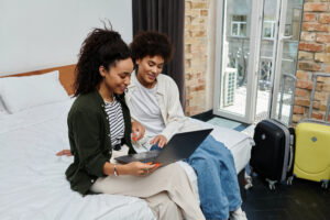 A couple seated on the edge of a bed, looking at a laptop computer screen together; traveling while on long-term disability requires careful preparation.