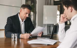 An attorney and disabled client seated at a conference table, reviewing a multi-page document containing buyout terms with the help of a laptop computer and a pen for markup; discussing average LTD payout amounts to reach realistic LTD settlement value estimates.