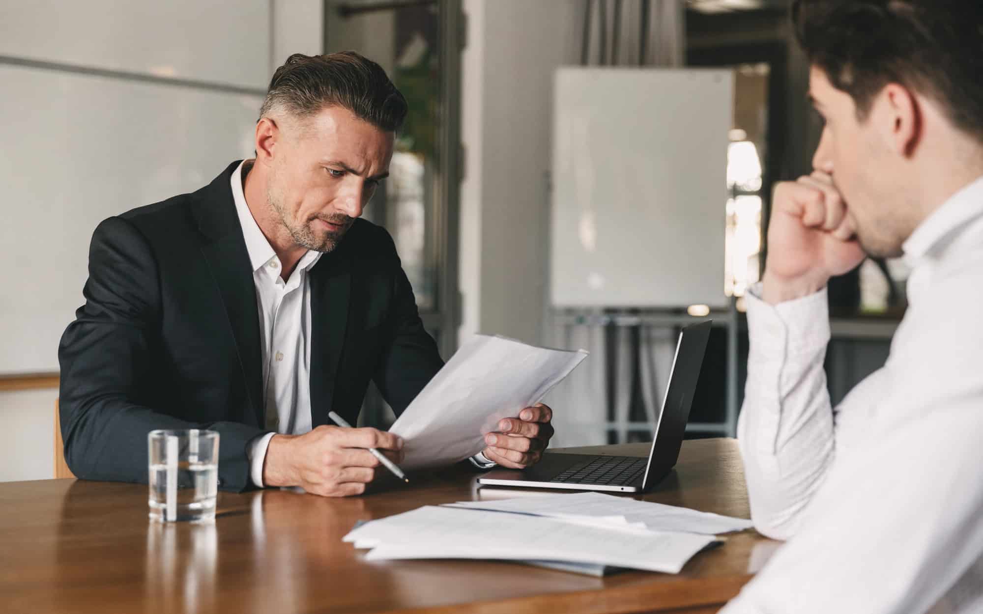 An attorney and disabled client seated at a conference table, reviewing a multi-page document containing buyout terms with the help of a laptop computer and a pen for markup; discussing average LTD payout amounts to reach realistic LTD settlement value estimates.