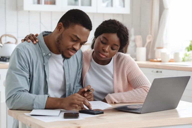 A man and woman sit together at a kitchen table spread with policy documents and an open laptop computer, calculating LTD impact on retirement savings with the assistance of a handheld calculator.