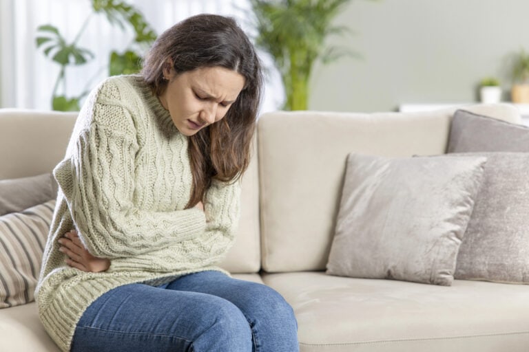 Young woman sitting on couch hugging her abdomen, grimacing in pain; health disruptions may trigger long-term disability insurance claims.