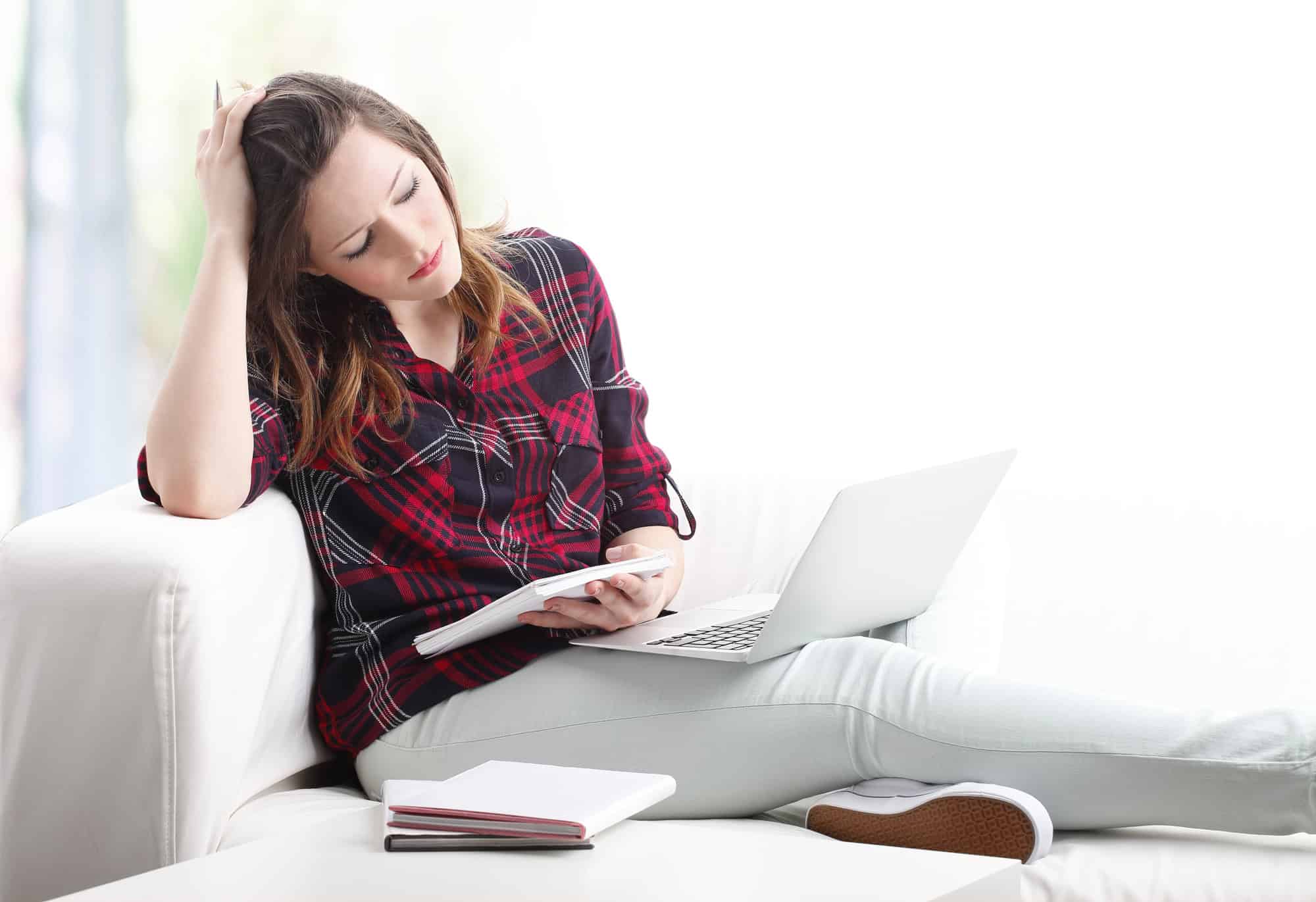 Exhausted young woman seated on sofa, reviewing handwritten notes with a laptop open on her legs; understanding common reasons LTD claims are denied.