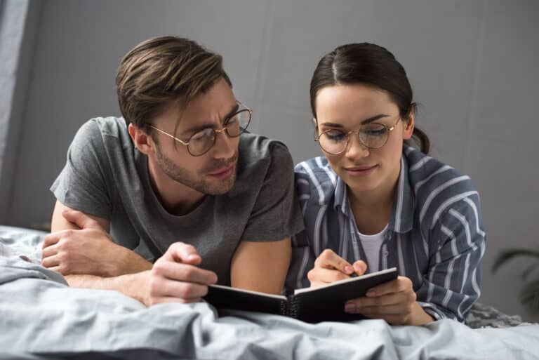 A married couple propped on their elbows in bed reviewing a notebook together; LTD benefits reduced reasons actions for family budgets.