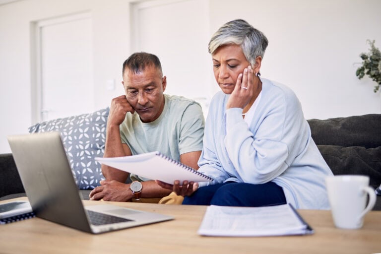 Senior couple sitting on a sofa in front of a coffee table, reviewing retirement planning documents together with an open laptop computer and three spiral-bound booklets; transitioning from LTD to retirement benefits.