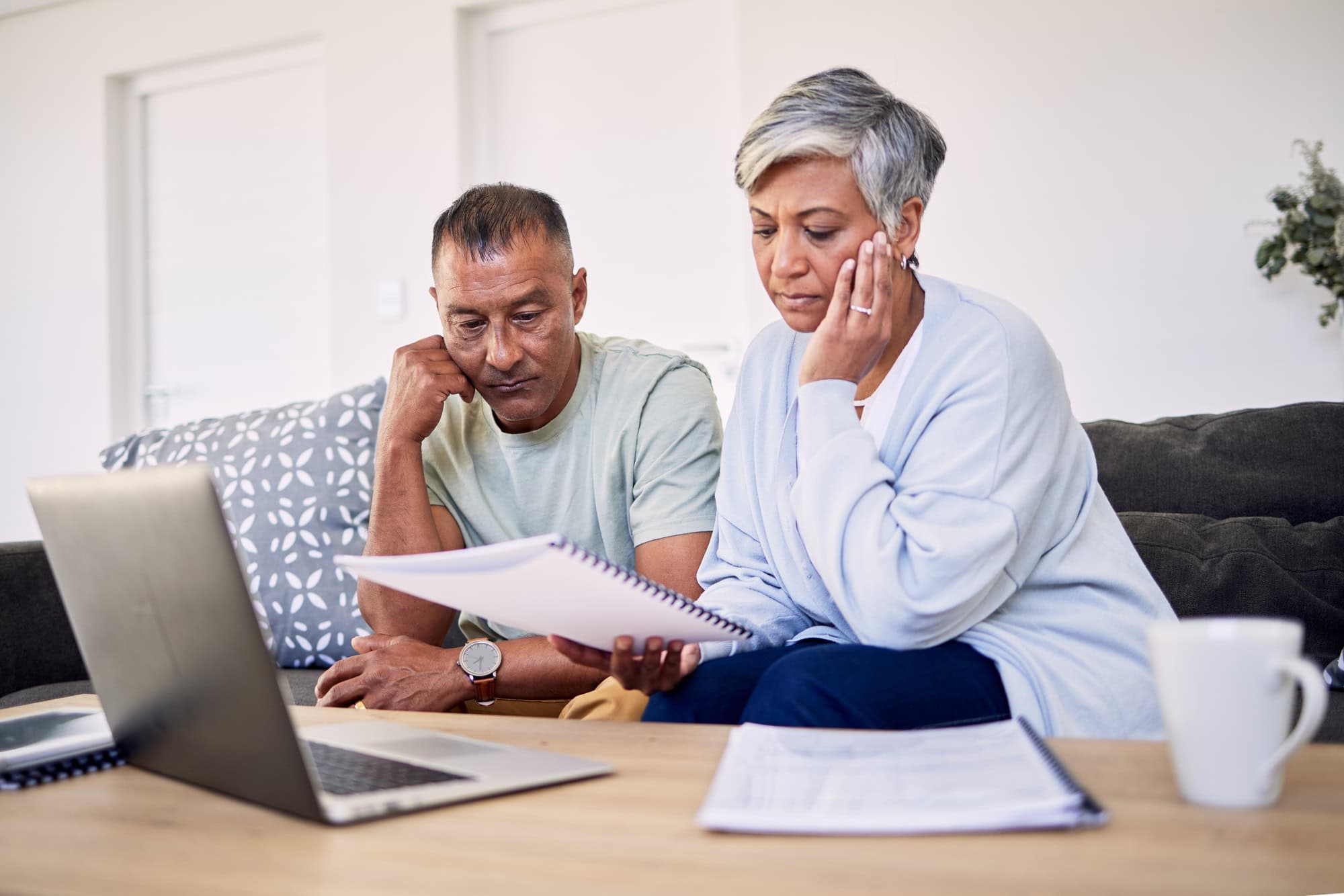 Senior couple sitting on a sofa in front of a coffee table, reviewing retirement planning documents together with an open laptop computer and three spiral-bound booklets; transitioning from LTD to retirement benefits.
