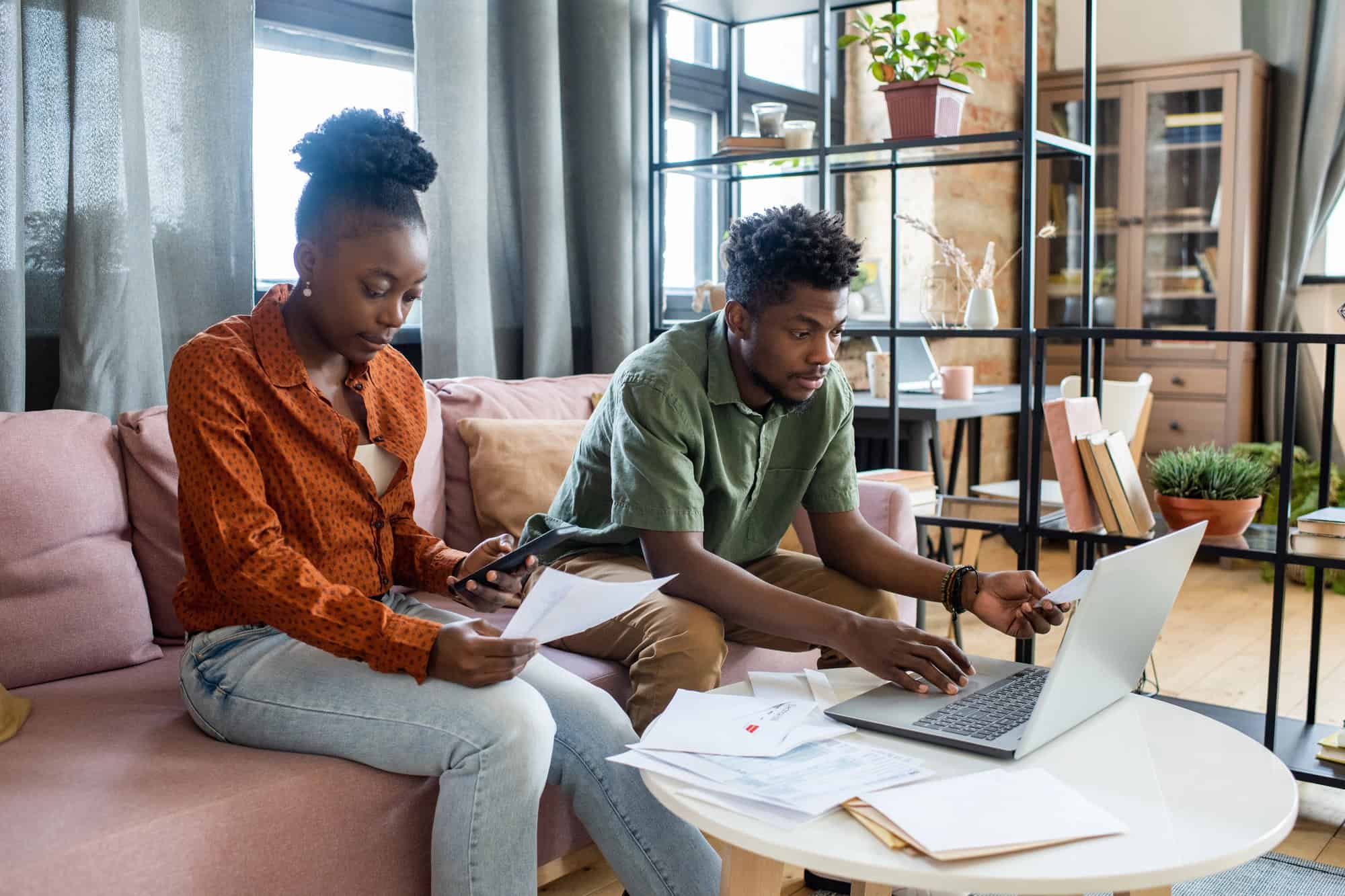 A young couple is sitting together on a sofa in front of a coffee table in an open plan apartment; the coffee table is covered in printed documents and manila folders, with a laptop computer open on one side. One partner works on the computer while the other calculates bills from a paper held in one hand using her phone. The absence of LTD benefits inflation adjustment (COLA) can complicate financial planning strategies.