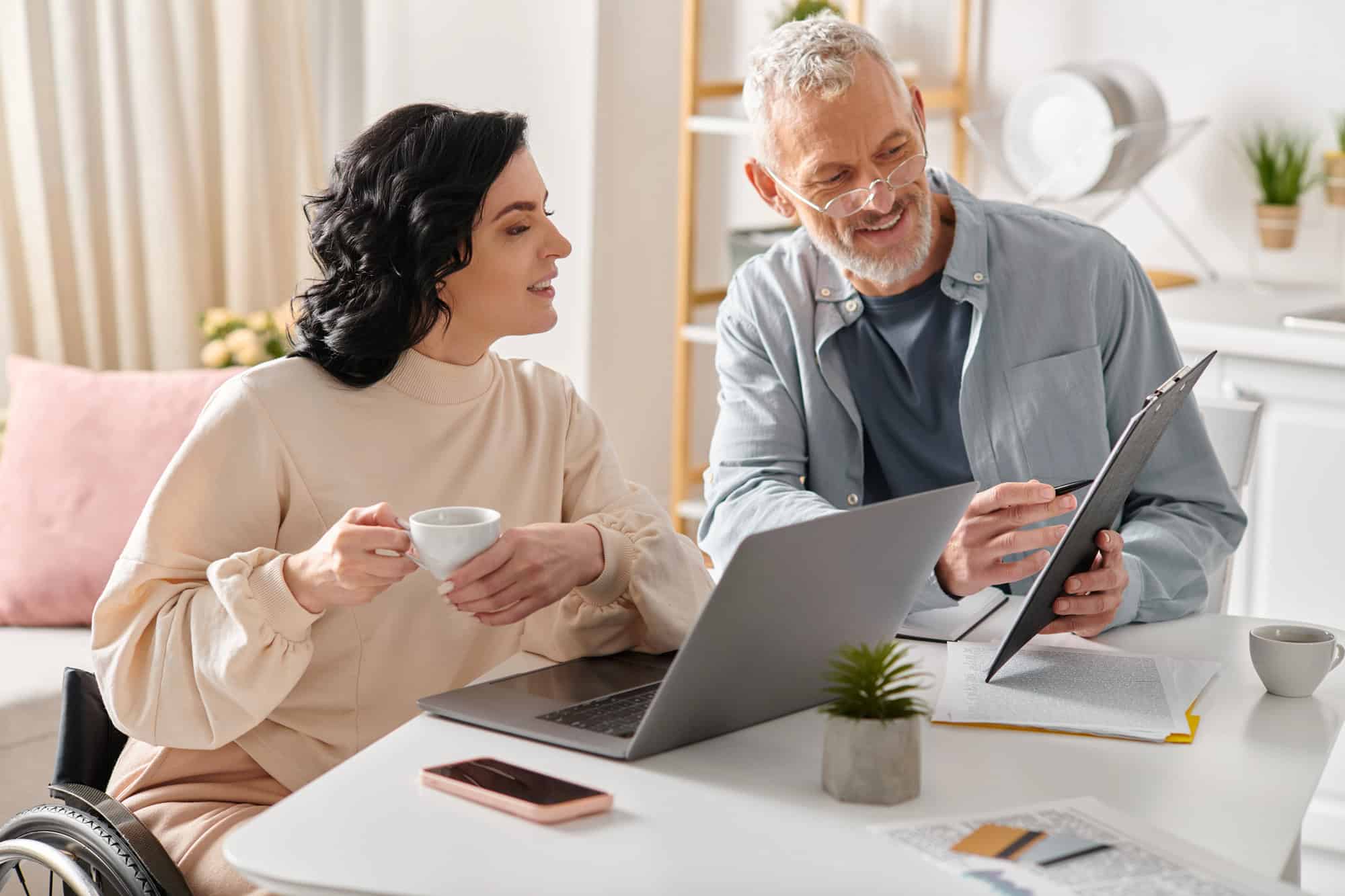 A woman in a wheelchair sitting at a kitchen table with a laptop and cup of coffee, reviewing documents with the assistance of an older man holding up a clipboard; getting help from an LTD benefits renewal process guide.
