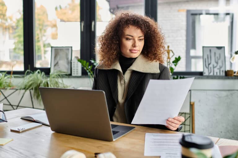 A young woman sitting at a desk with a laptop holds up a paper; reviewing policy terms to decide, “Is LTD a good investment?”