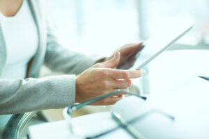 Closeup of a woman holding a tablet at a desk with a pair of glasses laid across an open notebook to one side; reviewing long-term disability options if employer offers no LTD.