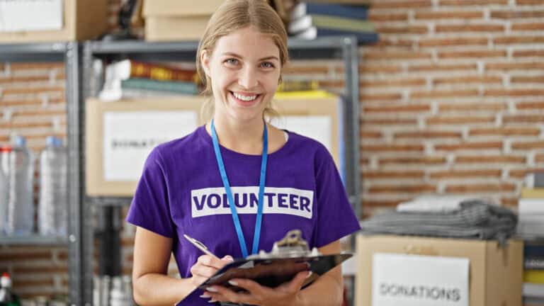 A young woman in a purple T-shirt with a strip of white background cut-out text that says “VOLUNTEER” across the front smiles at the camera as she uses a pen to mark off items on a clipboard; managing volunteer work impact on LTD benefits.
