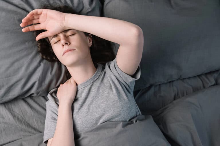 ME/CFS patient lying on her back in gray sheets, with one hand resting against her forehead and the other gripping her neck; navigating chronic fatigue syndrome (CFS) in LTD policies can prove difficult when brain fog and overwhelming exhaustion interfere with the ability to function.