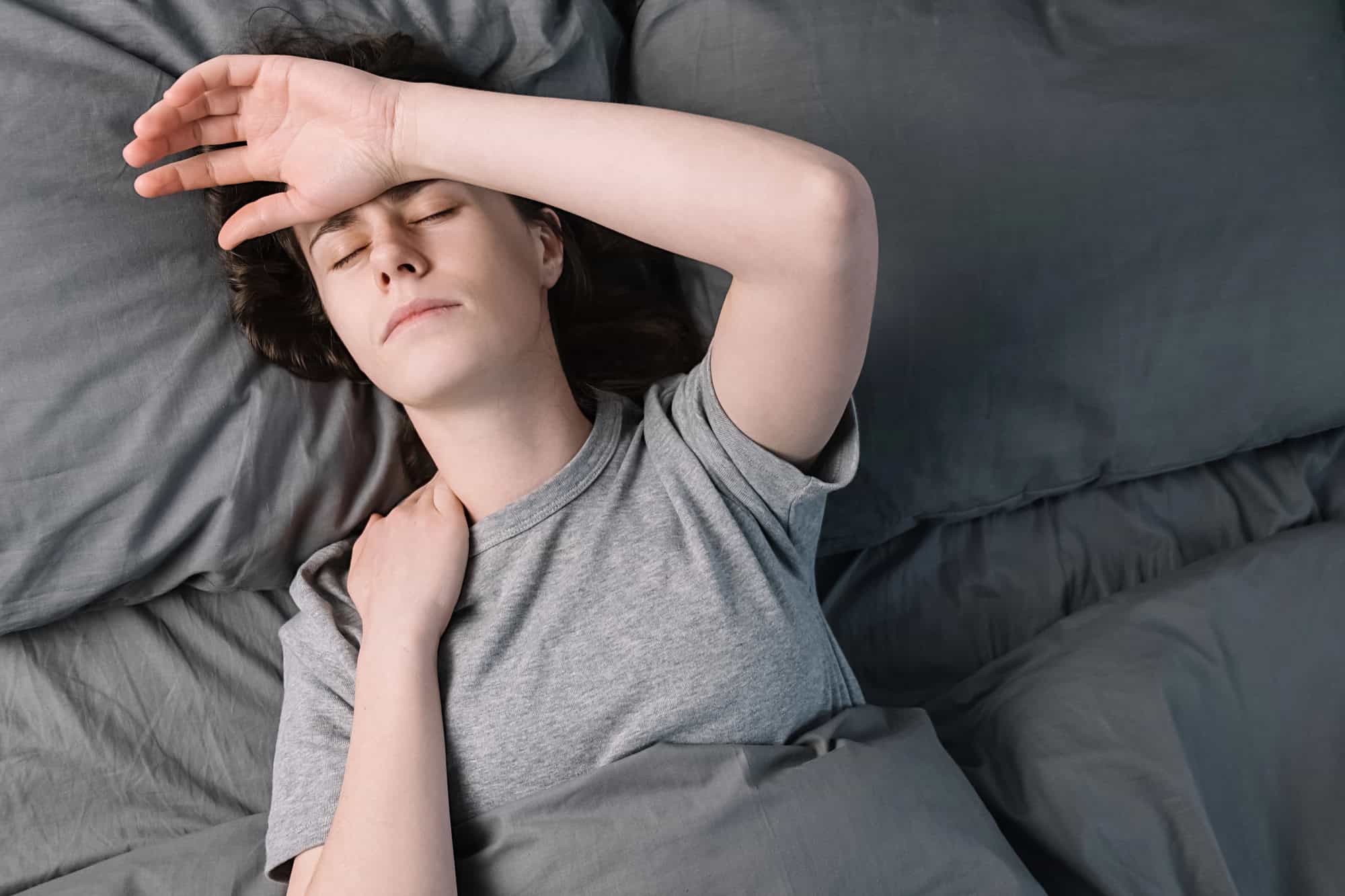 ME/CFS patient lying on her back in gray sheets, with one hand resting against her forehead and the other gripping her neck; navigating chronic fatigue syndrome (CFS) in LTD policies can prove difficult when brain fog and overwhelming exhaustion interfere with the ability to function.