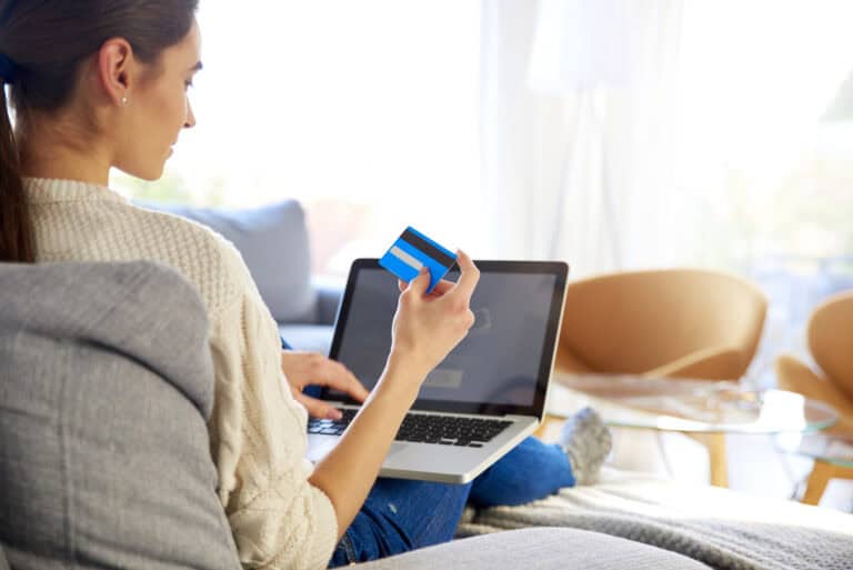 Over-the-shoulder view of a woman sitting on a sofa and holding up a credit card in one hand while the other taps the keys of a laptop computer; reconsidering online purchases in light of LTD impact on credit scores assessments.