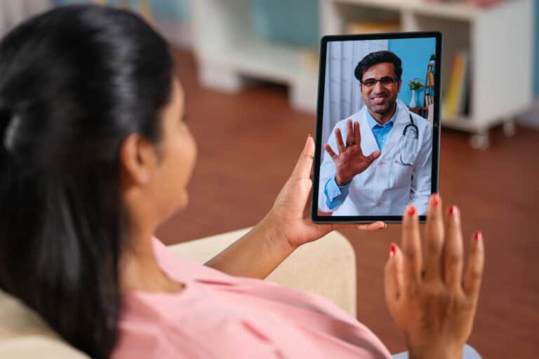 Woman leaning back into an armchair, consulting with a medical doctor via a tablet held in one hand; navigating LTD benefits and telehealth services.