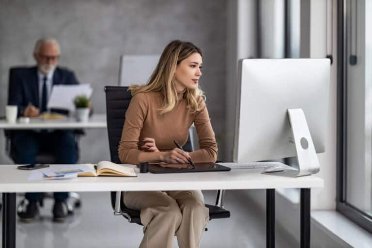 A woman in business casual attire is seated at a desk in an open plan office, taking notes in front of a desktop computer with documents spread across the desk. Balancing part-time work and LTD can be fulfilling when managed carefully.