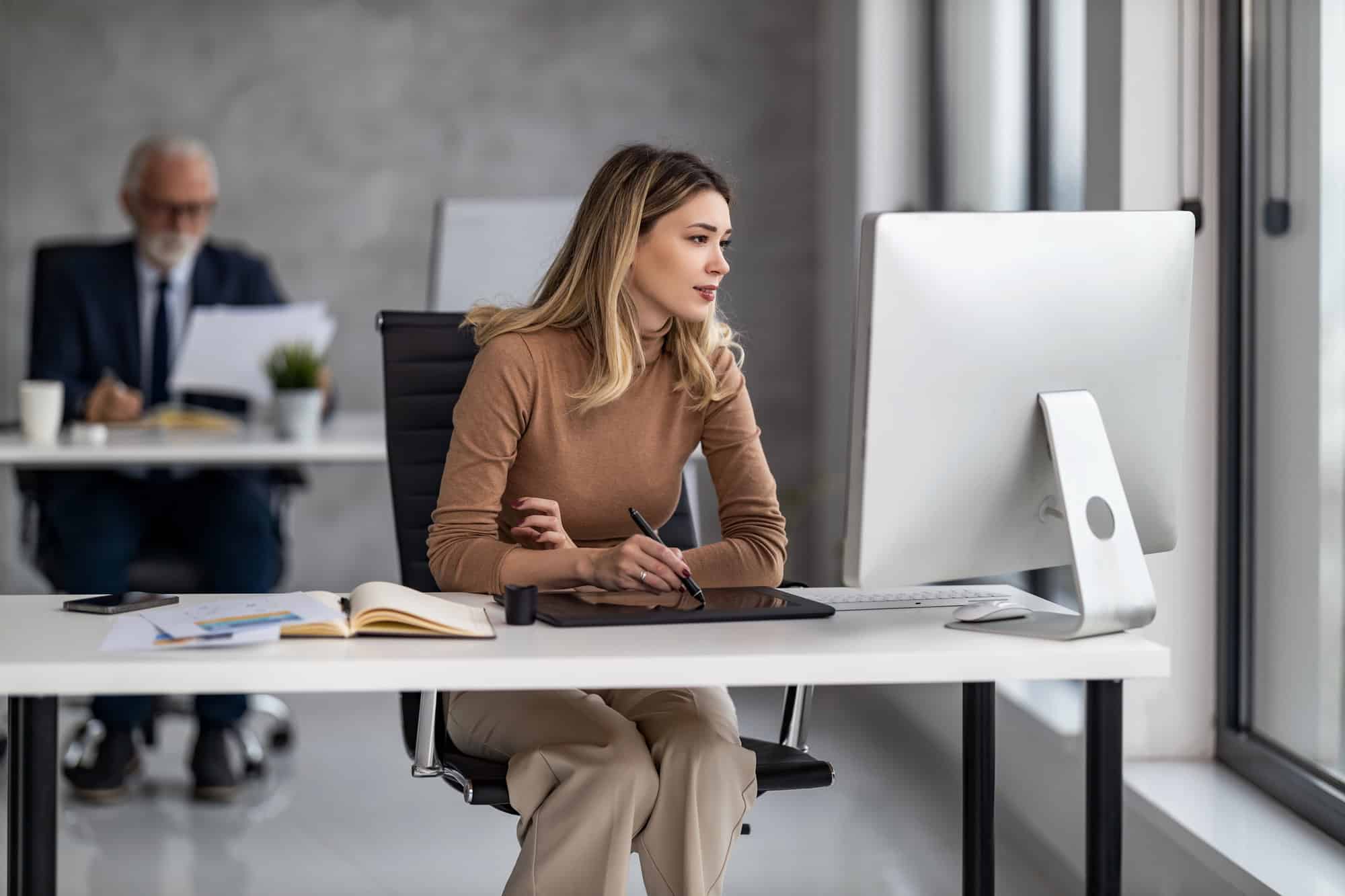 A woman in business casual attire is seated at a desk in an open plan office, taking notes in front of a desktop computer with documents spread across the desk. Balancing part-time work and LTD can be fulfilling when managed carefully.