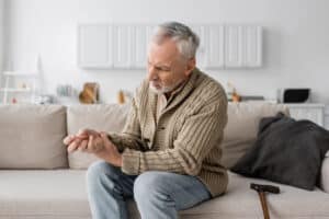 LTD for progressive medical conditions needs to account for gradual increase in impairments and a man with gray hair seated on a sofa next to a walking cane, attempting to uncurl the fingers of one