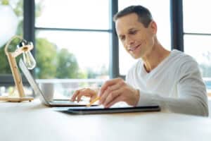 Middle-aged man seated at a desk, using a set of paper documents and a laptop computer to conduct a short-term vs. long-term disability insurance comparison.