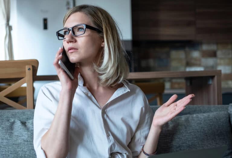 A woman in glasses and a button-down shirt having a phone conversation seated on a sofa in a domestic setting; negotiating LTD insurance terms guide to success.