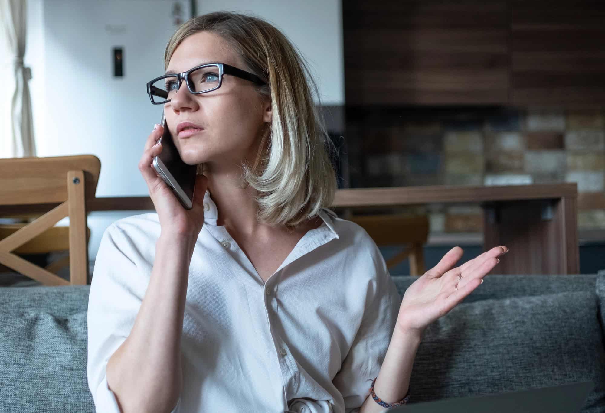 A woman in glasses and a button-down shirt having a phone conversation seated on a sofa in a domestic setting; negotiating LTD insurance terms guide to success.