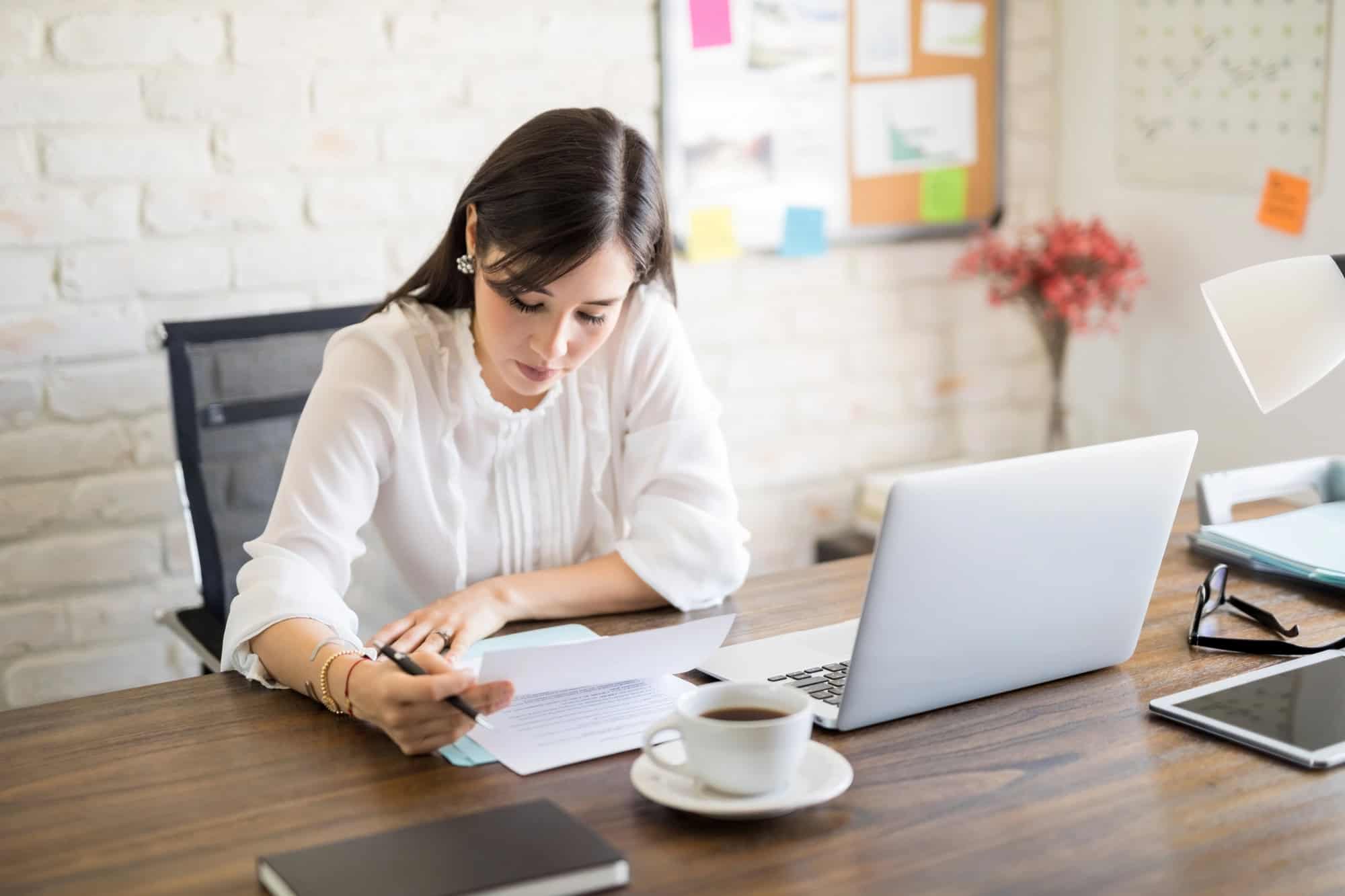 A young woman in a home office setting reviewing printed policy documents at a desk with a cup of coffee and a laptop computer; selecting best LTD insurance policy options.