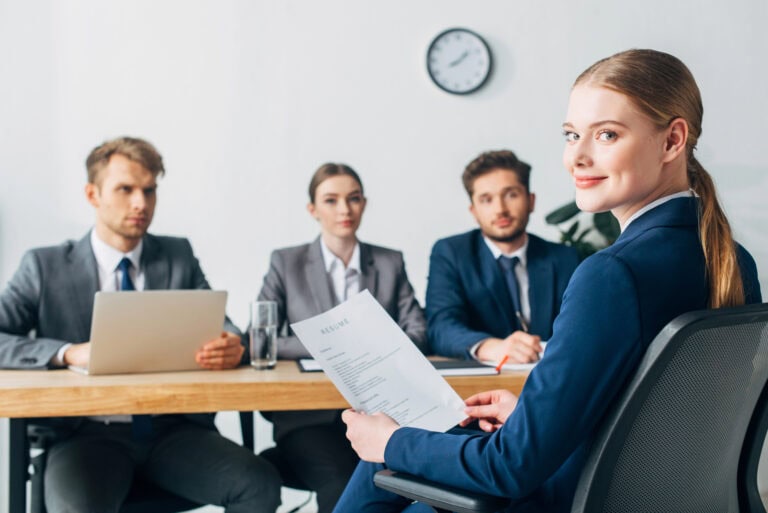 A female job candidate seated in front of a row of male and female interviewers glances back over her shoulder to smile at the camera; navigating LTD impact on future job prospects.