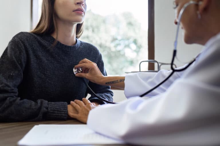 A doctor in a white lab coat listens to a female patient’s heartbeat in a medical exam room; pre-existing conditions and LTD eligibility overview often relies heavily on an individual’s history of medical care.