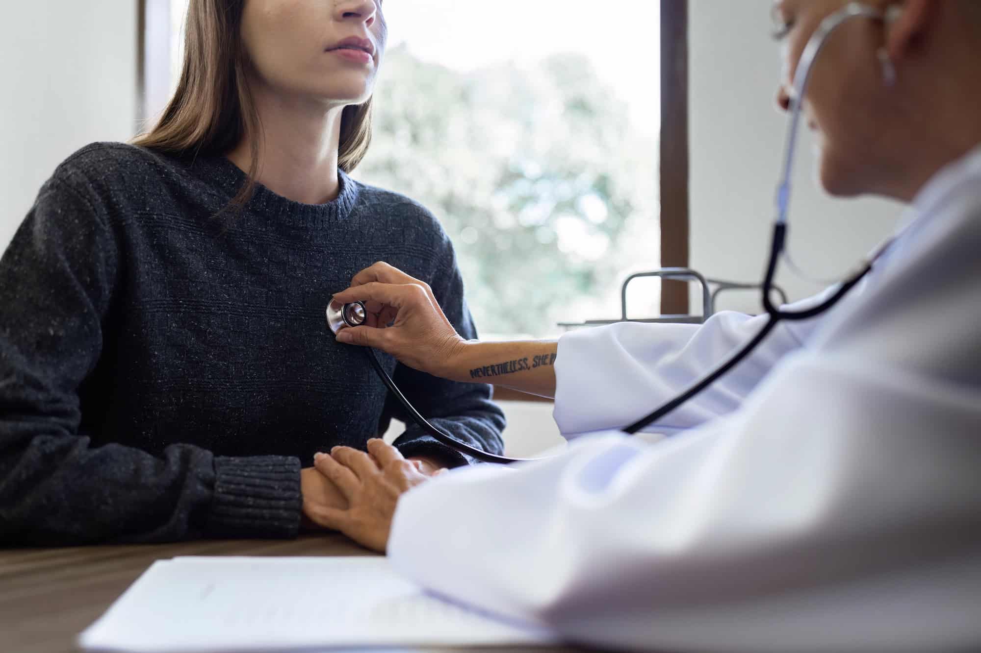 A doctor in a white lab coat listens to a female patient’s heartbeat in a medical exam room; pre-existing conditions and LTD eligibility overview often relies heavily on an individual’s history of medical care.
