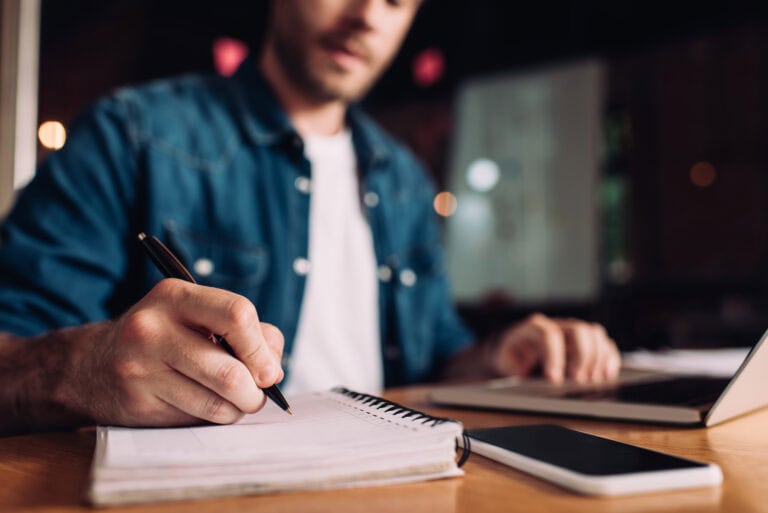 A young man in a white T-shirt under an open denim button-down taking notes in a spiral-bound notebook while reviewing information on a laptop computer; preparation for navigating LTD application pitfalls.