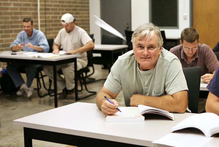 A middle-aged man looks up from his textbook at a desk shared with a partner in a classroom filled with younger adults; educational funding for LTD recipients helps individuals on long-term disability retrain for alternative career paths.