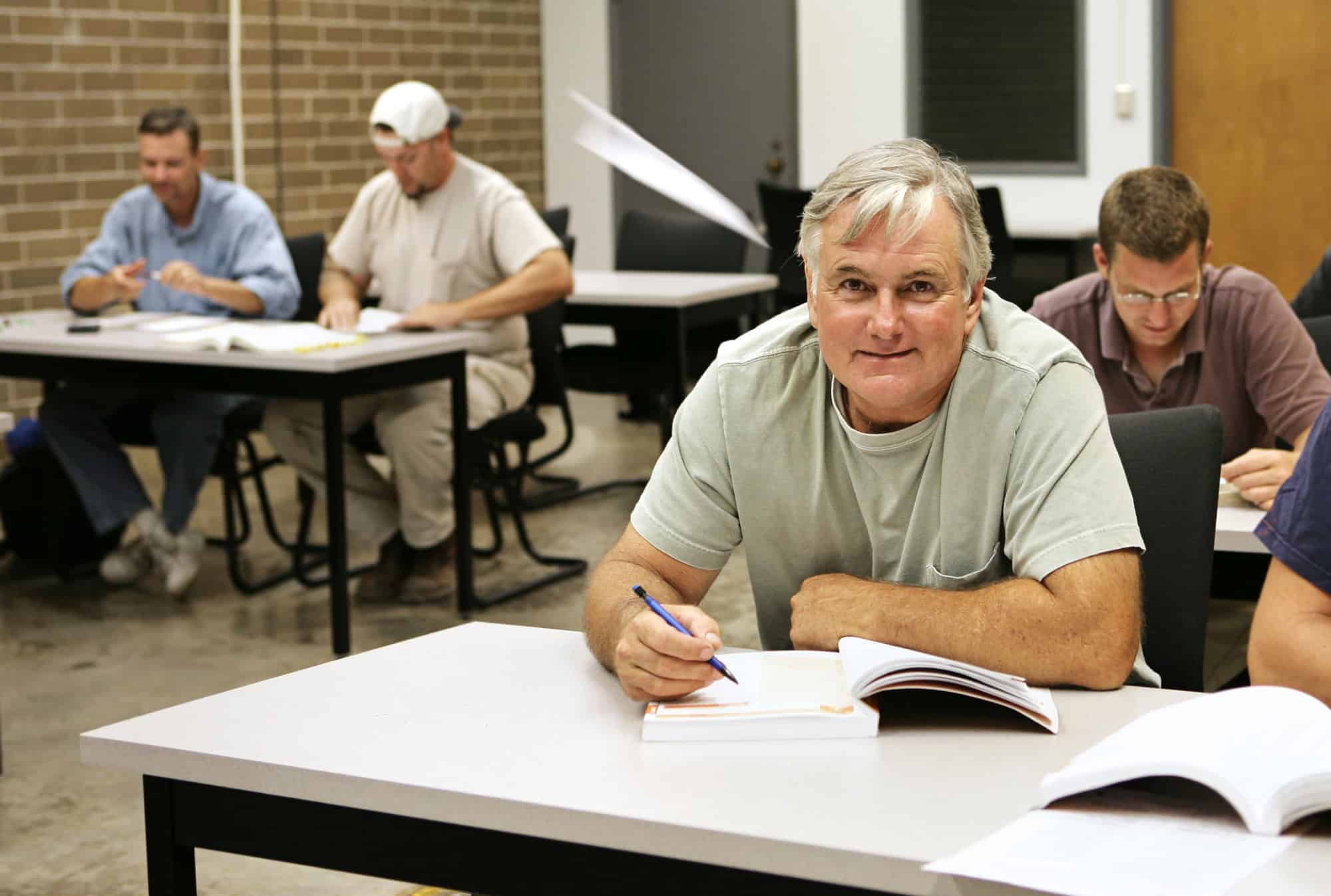 A middle-aged man looks up from his textbook at a desk shared with a partner in a classroom filled with younger adults; educational funding for LTD recipients helps individuals on long-term disability retrain for alternative career paths.