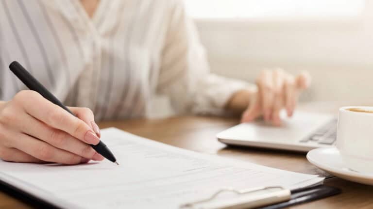 Closeup of a woman taking notes on a clipboard while reviewing information on a laptop computer, a cup of coffee sitting between computer and clipboard; taking the first steps toward navigating LTD benefits with other income sources.