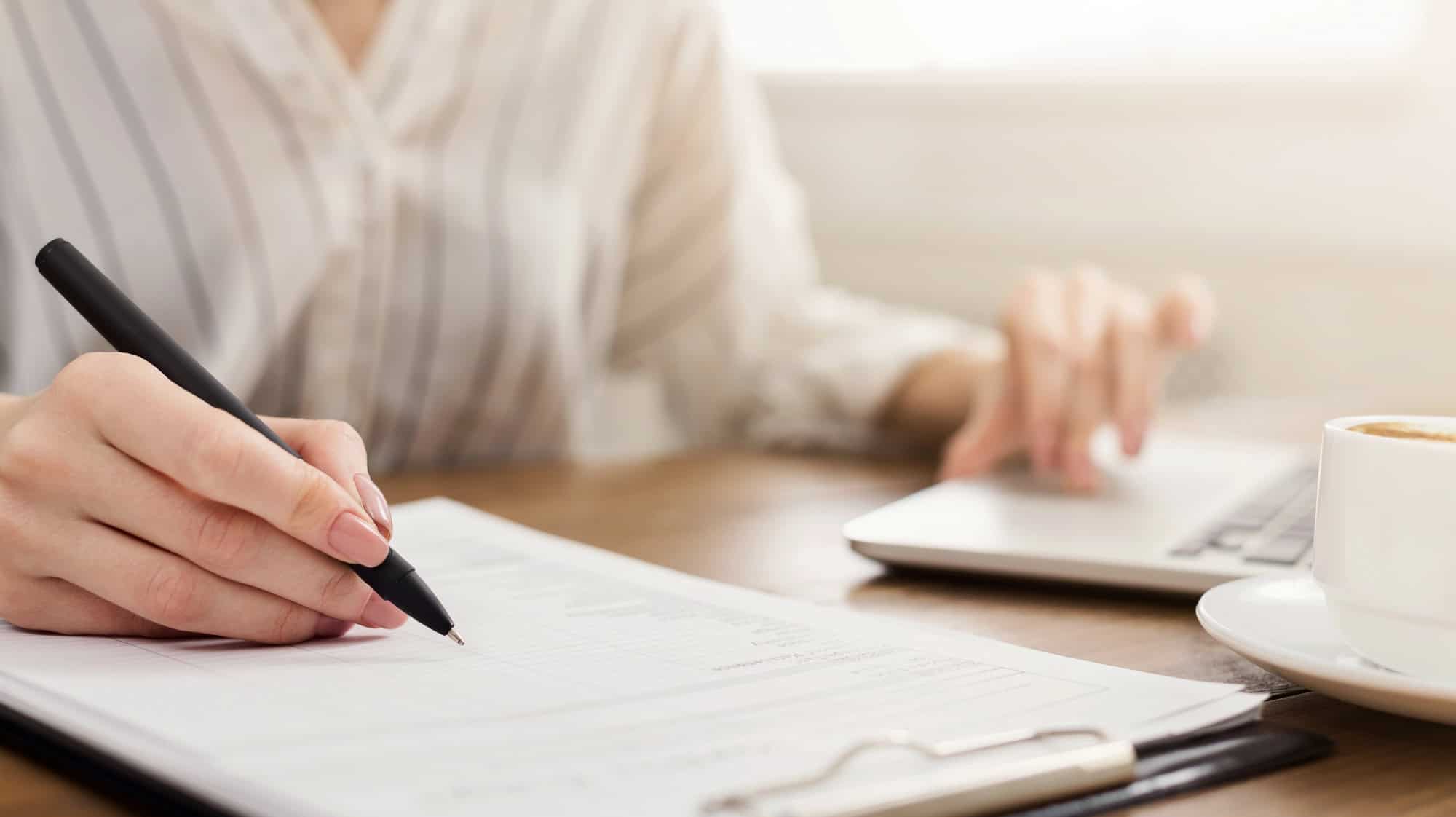 Closeup of a woman taking notes on a clipboard while reviewing information on a laptop computer, a cup of coffee sitting between computer and clipboard; taking the first steps toward navigating LTD benefits with other income sources.