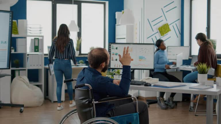 A middle-aged man in a wheelchair raises his hand during a group brainstorming session in a modern office space with a multicultural group of team members; accommodations can be essential to protecting LTD rights in the workplace.