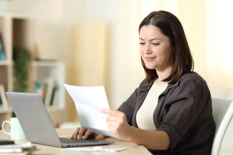 A woman sitting at a table in a domestic environment, reading a letter held in her left hand while navigating the trackpad on a laptop with her right; coordinating LTD with other personal insurance effectively can improve peace of mind.