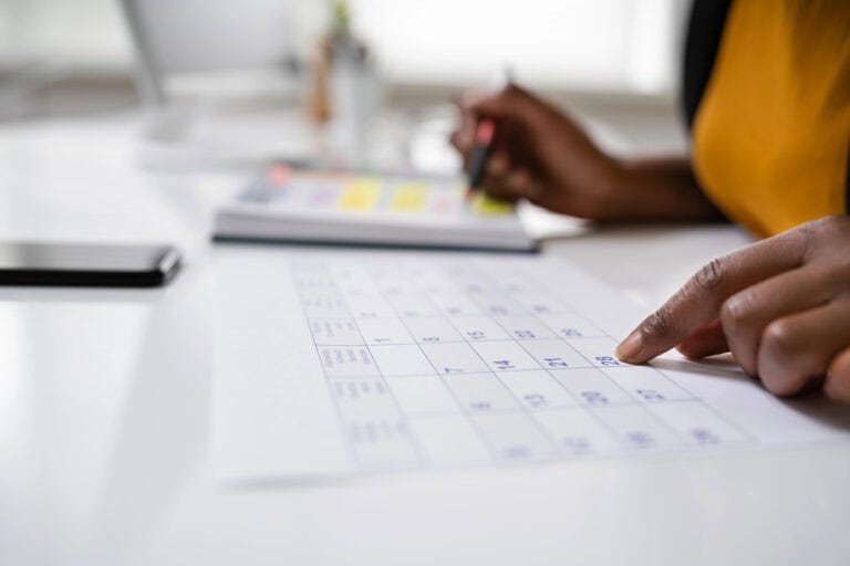 Closeup of a desktop with a woman’s left hand resting one fingertip on a desk calendar and her right hand poised with a pen above an agenda book, while a smartphone sits on the desk in front of the calendar; how technology helps you manage your long-term disability claim.