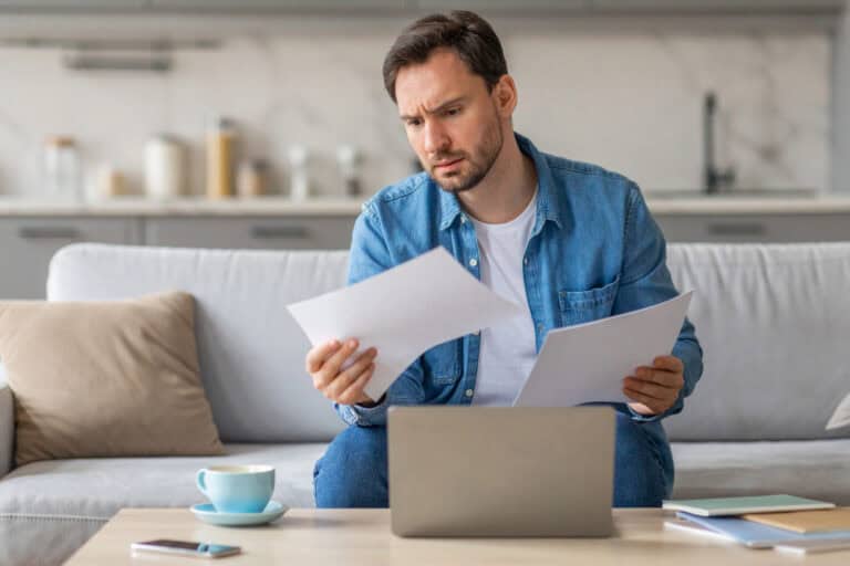 A man in casual attire sitting on a light-colored sofa reviewing a printed document in each hand in front of a coffee table holding a laptop computer, a phone, a cup of coffee, and several books or handbooks; group vs. individual LTD policies compared at home.