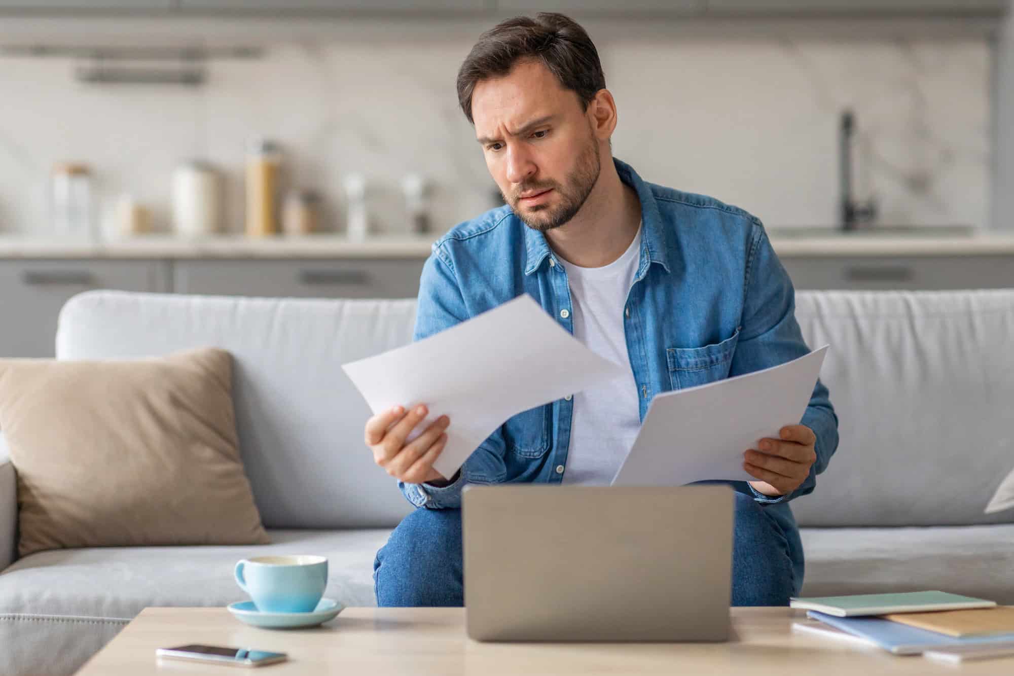 A man in casual attire sitting on a light-colored sofa reviewing a printed document in each hand in front of a coffee table holding a laptop computer, a phone, a cup of coffee, and several books or handbooks; group vs. individual LTD policies compared at home.