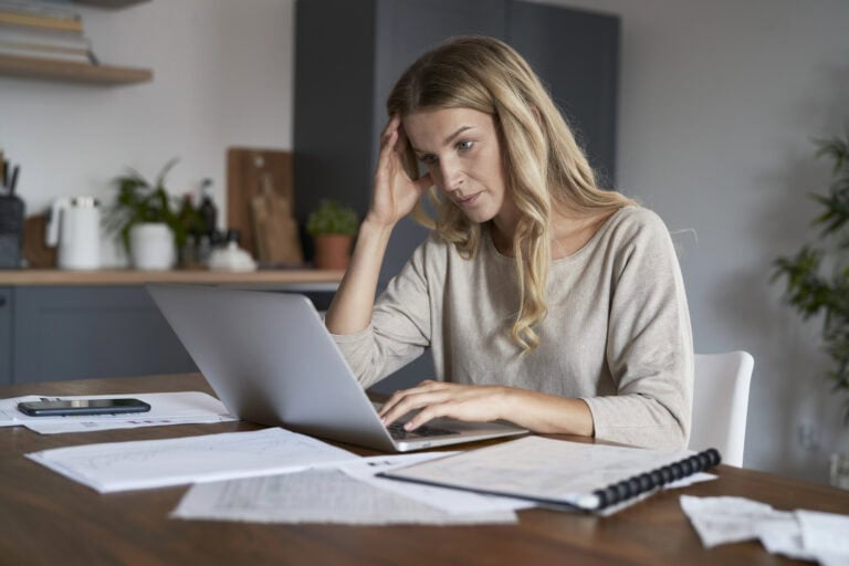 A woman sits at a desk, working on a laptop computer. The desk is strewn with printed documents, a binder, and a mobile phone, as the woman searches for a step-by-step LTD application guide.