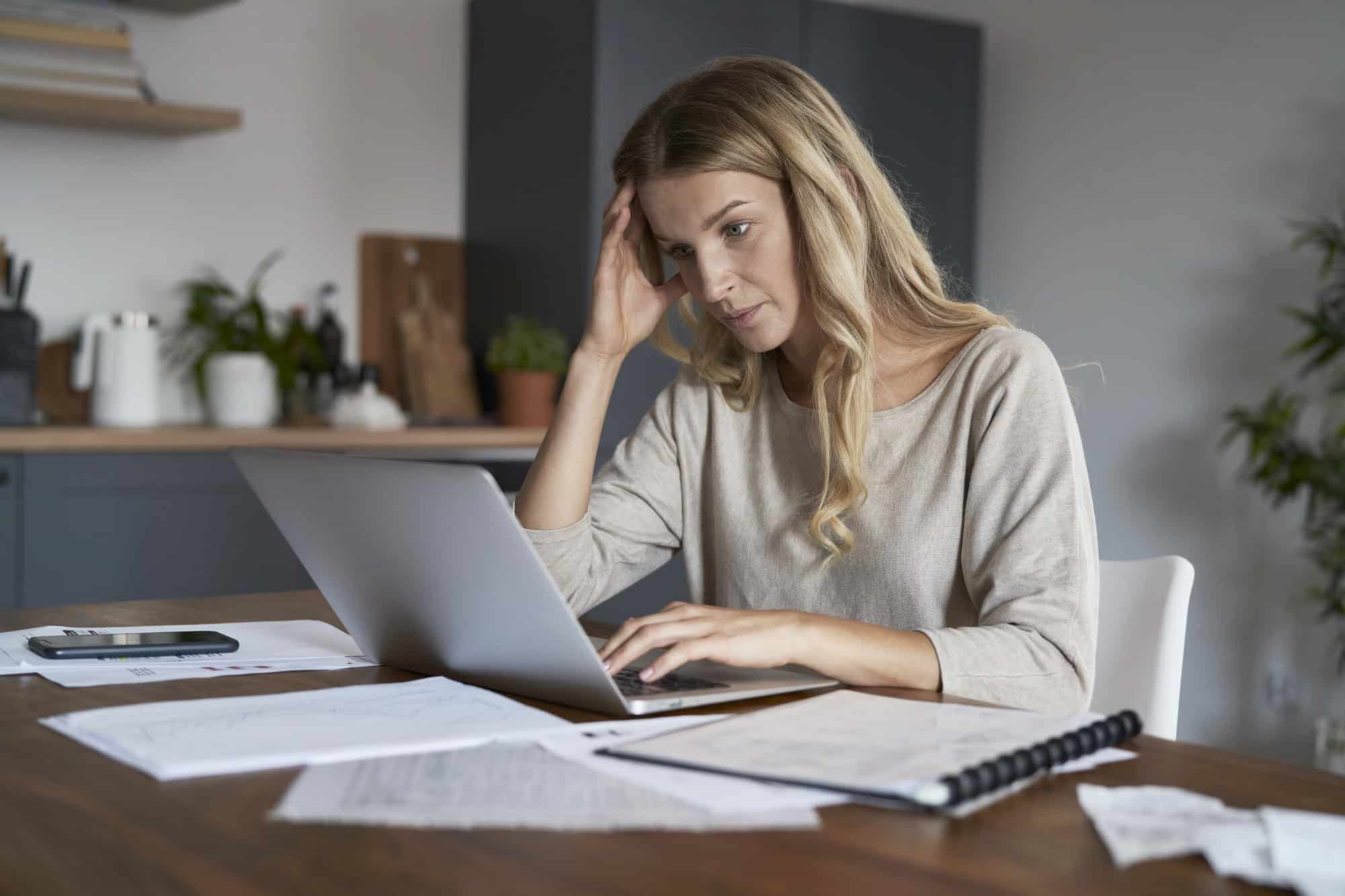 A woman sits at a desk, working on a laptop computer. The desk is strewn with printed documents, a binder, and a mobile phone, as the woman searches for a step-by-step LTD application guide.